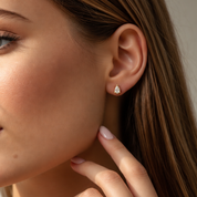 A close-up of a woman's ear wearing a 1.00ct pear-cut diamond stud earring in a gold setting, with her hand resting near her neck.