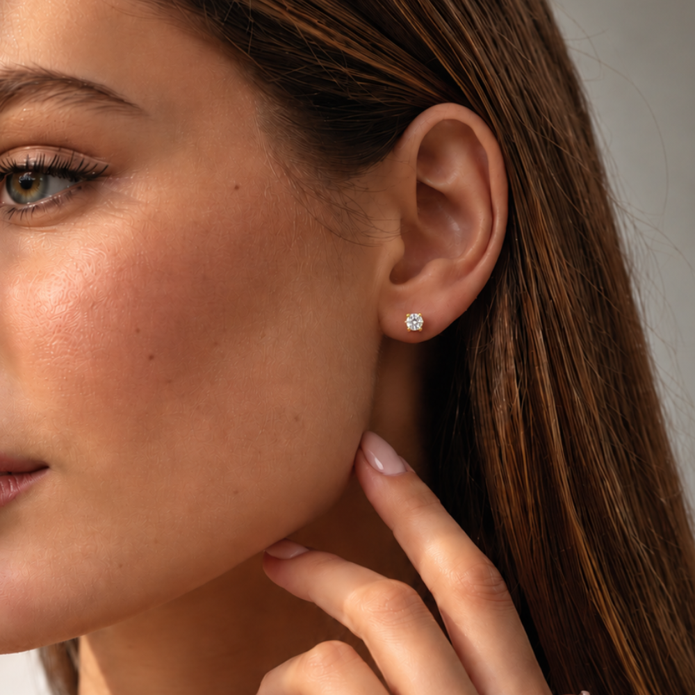 A close-up of a woman's ear wearing a small round diamond stud earring. The earring is set in a yellow gold metal and is shown on a model with brown hair and light skin against a soft gray background.