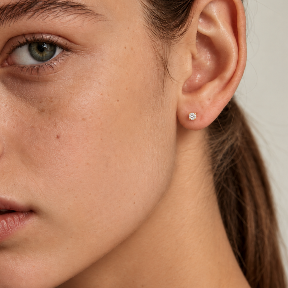 A close-up of a woman's ear wearing a small round stud earring with a lab-grown diamond in 14k yellow gold