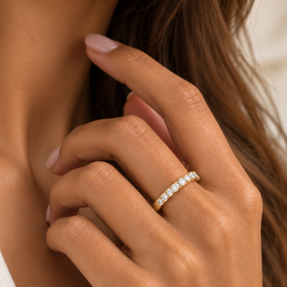 A close-up lifestyle shot of a woman's hand wearing a yellow gold half-eternity ring set with small round diamonds. The ring is shown on her ring finger against a soft-focus background of her skin and brown hair.