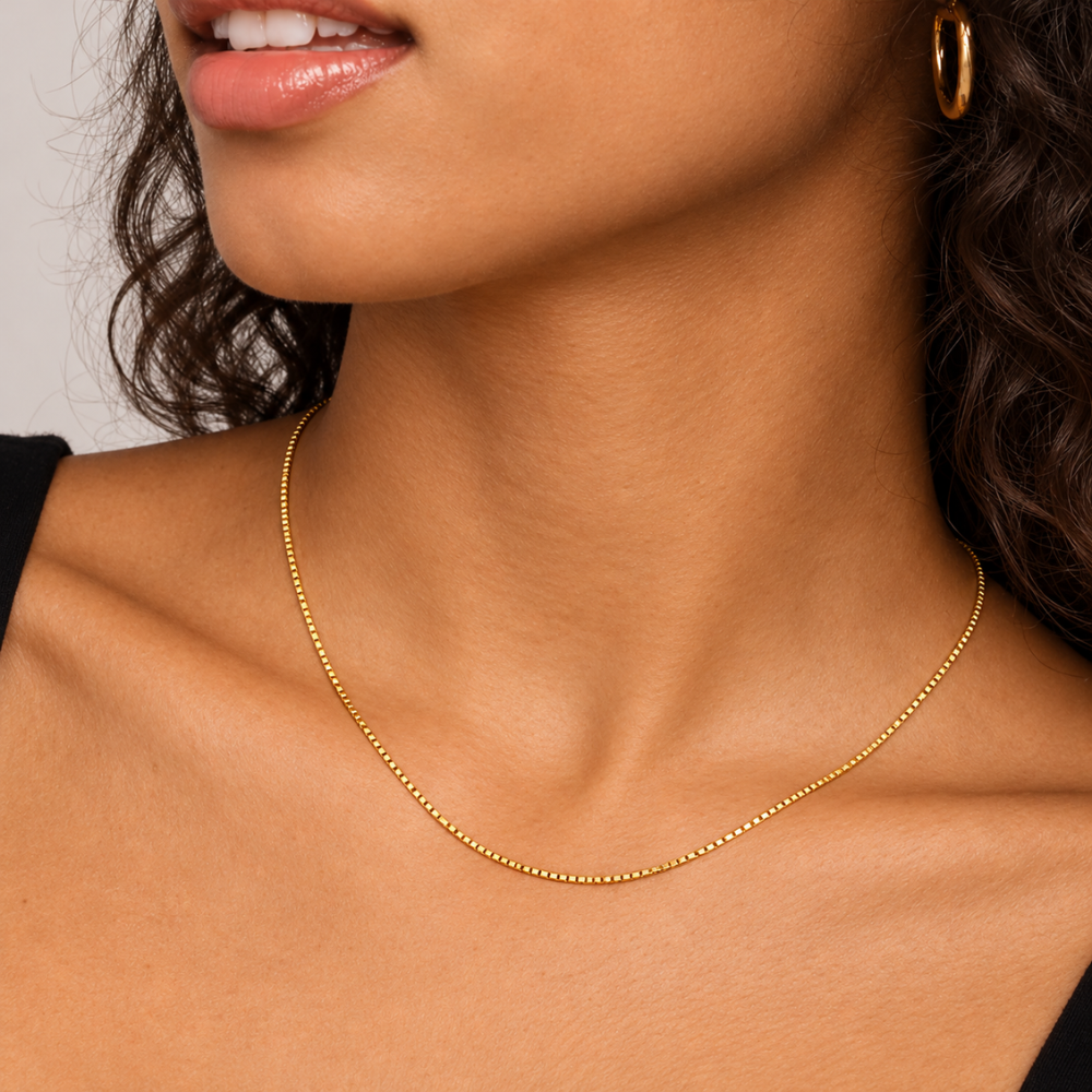 A close-up of a model wearing a delicate gold chain necklace and a gold hoop earring, showing the jewelry on the neck.