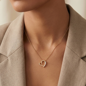 A close-up of a model wearing a delicate gold heart-shaped pendant necklace with small clear stones, styled with a beige blazer.
